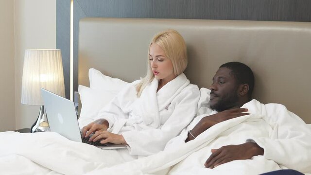 Mixed race couple on bed in hotel, woman work on laptop, african boyfriend sits next to her, support.