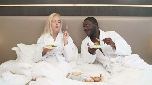 Adorable multiethnic couple have breakfast in hotel, they have tasty meal together, enjoy, relax, happy weekends. on bed.