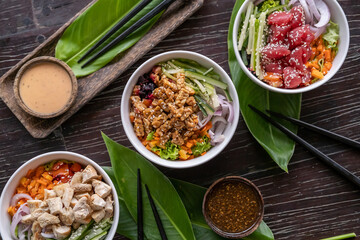 Variety of healthy organic poke bowls with tuna, chicken, tofu and cucumber, rice, carrots, onion served on wooden table with tropical leaves. Healthy lifestyle