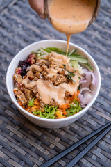 Close up of a hand adding dressing to healthy organic vegetarian poke bowl with tofu, fermented soybeans -tempeh, cucumber, rice, carrots, onion served in bowl on table. Healthy lifestyle