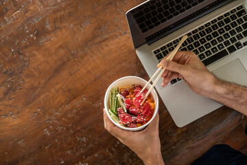 Busy young man in casual wear sitting at desk with a laptop at home or in the office and eating healthy takeout poke bowl food from container during lunch break. Healthy lifestyle concept. Top view - Powered by Adobe