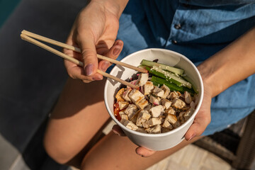 High angle of woman sitting with chopsticks and eating takeaway healthy organic chicken poke bowl...