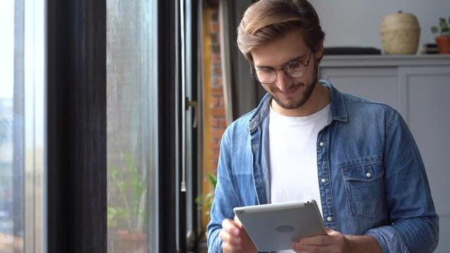 Young professional man worker using digital tablet standing at home office, smiling male manager working online on pad computer tech