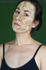  young brunette woman with a clay homemade mask on her face looks into the frame