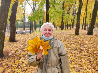 Older woman in protective mask and a bouquet of autumn leaves is having fun in the autumn park outdoors.