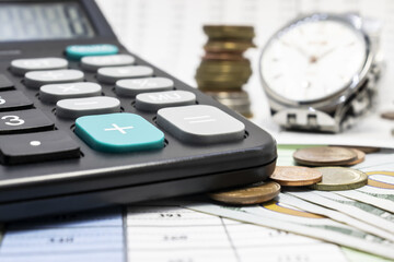On the document with money, a black calculator is close-up on a blurry background of a clock and a stack of coins. Business concept