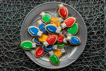 Christmas lightbulb cookies with string on a silver platter against a silver textured background.