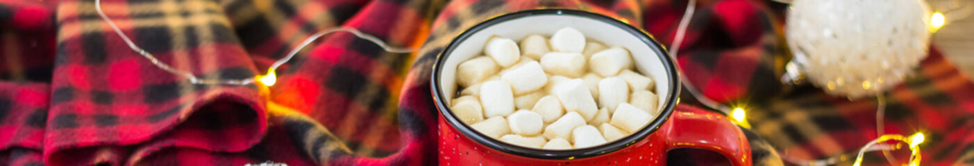 Banner of Christmas coffee in a red cup with marshmallows and plaid. Decorated white balls and light garland bokeh