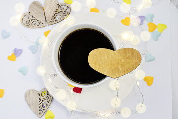 Against the background of colorful hearts with bokeh is a white Cup of coffee on which there is a wooden heart with space for text. Holiday background
