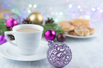 Obraz na płótnie Canvas Christmas background with a purple ball on the foreground and a blurred image of a cup of coffee with milk, cookies, sprigs of fir and Christmas balls.