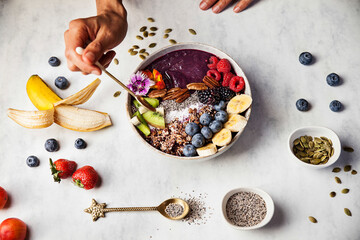 A woman is eating an açai bowl with fresh fruits for breakfast with banana, blueberries, blackberries, strawberries, kiwi, nuts and seeds.