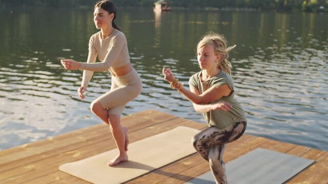 Two young caucasian women in sportswear practicing eagle pose while doing yoga together on wooden pier beside mountain lake