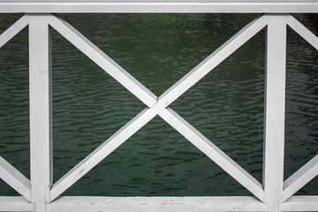 View of the reservoir through the embankment fencing elements. White painted wood fencing parts. Vertical and diagonal fence elements on a sunny day. Side view.