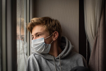 Young ill teenage boy looking sad with protective mask at home behind window in quarantine and lockdown missing school and freedom during Covid-19 Coronavirus worldwide pandemic