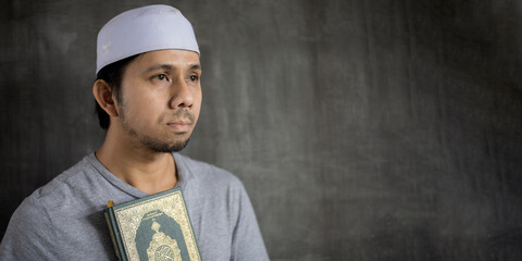Portrait of a young muslim man Holding the book of the holy quran against grey wall background with...