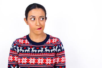 Amazed puzzled Young beautiful Arab woman wearing Christmas sweater against white wall, curves lips and has worried look, sees something awful in front.