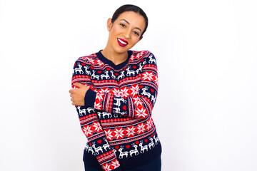 People, lifestyle, youth and happiness concept. Shy pretty Young beautiful Arab woman wearing Christmas sweater against white wall, feeling happy hugging herself.