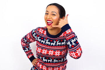 Young beautiful Arab woman wearing Christmas sweater against white wall imitates telephone conversation, makes phone call gesture with hands, has confident expression. Call me!