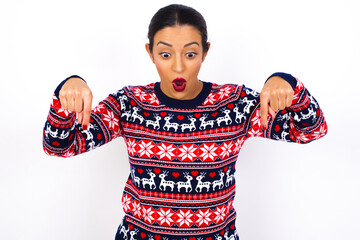 Amazed Young beautiful Arab woman wearing Christmas sweater against white wall points down with...
