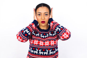 Fototapeta premium Frustrated Young beautiful Arab woman wearing Christmas sweater against white wall, plugging ears with hands does not wanting to listen hard rock, noise or loud music.