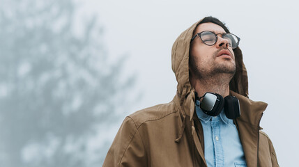 Attractive male walking in misty nature, listening to the music on his headphones with closed eyes outside. Traveler man wearing a stylish coat in the mountain enjoying the fresh air.