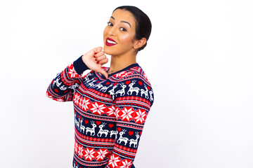 Closeup of cheerful Young beautiful Arab woman wearing Christmas sweater against white wall, looks joyful, satisfied and confident, points at himself with thumb.