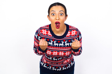 Joyful excited lucky Young beautiful Arab woman wearing Christmas sweater against white wall, cheering, celebrating success, screaming yes with clenched fists