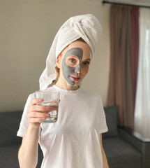 young woman in a towel on her head having a facial beauty Spa with a clay mask at home, she holds a glass of water.