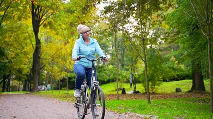happy senior woman in headphones rides park a bicycle and listens to music audio book. old female cyclist with a retro bike pursues active lifestyle in nature. walk autumn forest or city park outside