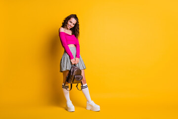 Full length photo of lady wavy hairdo toothy smile hold backpack wear pink top unclothed shoulders mini skirt white stockings sneakers isolated yellow color background