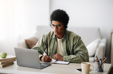 Industrious black teenager in headset studying remotely from home, writing in notebook