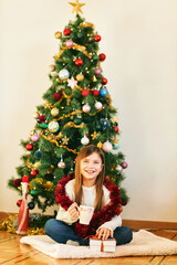 Portrait of sweet little girl sitting next to Christmas tree, wearing white cozy pullover, holding cup with hot chocolate, winter holidays