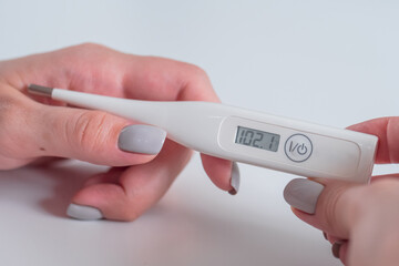 Woman hands holding white digital medical thermometer with high temperature - close up view,...
