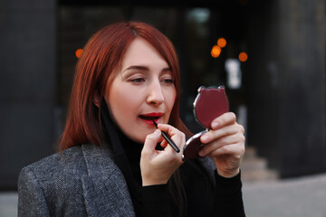 Redhead caucasian business woman looking at little pocket mirror and applying red lipstick after...
