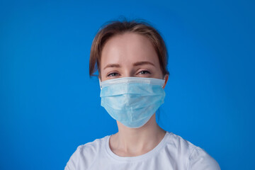 Portrait of woman wearing medical face mask, white shirt, looking at camera in room with blue wall at home - close up front view. Self isolation, quarantine, COVID19, coronavirus, safety concept