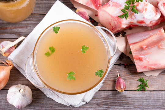Bone broth, bone stock in a glass, garlic cloves and herbs on the table, paleo diet. Top view