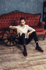 Fototapeta na wymiar A young man sits on the floor, leaning on a souvenir wooden clock. Trendy male with modern beard and strict outfit in the old antique apartment room photostudio. Vertical portrait, copy space. 