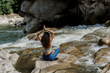 Yoga on the rocks near waterfall in mountain river. Girl is travelling in Karpathian mountains and feeling freedom. Cascade waterfall and beautiful young woman