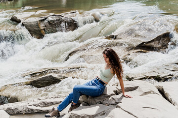 Traveler girl is sitting on the rock near waterfall and looking toward. Travelling in Karpathian mountains. Cascade waterfall. Beautiful landscape