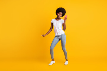 Fototapeta na wymiar Full length body size photo of beautiful young girl with black skin listening music dancing holding smartphone isolated on bright yellow color background