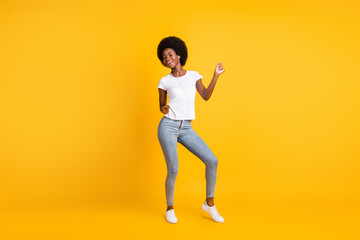 Full length body size photo of pretty active girl with black skin listening music dancing with earphones wearing t-shirt jeans smiling isolated on bright yellow color background