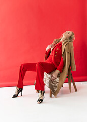 A beautiful blonde curly girl wearing a red pantsuit, unbuttoned on her braless big breasts, relaxes, sitting on a chair and closing her eyes on a red background. Advertising, fashionable design
