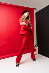 A beautiful blonde curly girl wearing a red pantsuit, unbuttoned on her braless big breasts and hat sensually hugs herself a red background. Advertising, fashionable, commercial design
