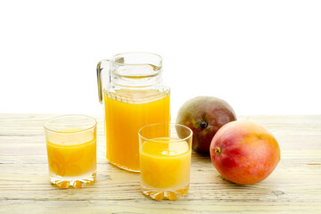 Tasty mango juice in a jug, glasses and ripe mango fruit on a wooden table close-up.