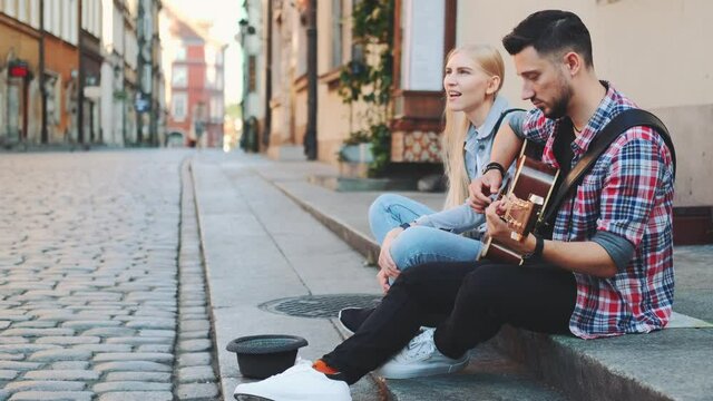 Young man and woman of street singers sitting on sidewalk, playing guitar and singing. Lifestyle concept.