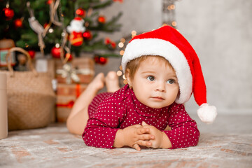 Cute baby Santa sits at home near the Christmas tree with gifts