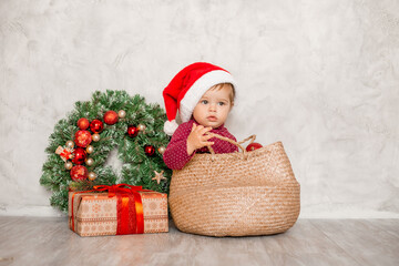 Sweet baby Santa sits in a wicker basket with a gift box and a Christmas wall on a gray background. Christmas concept, text space