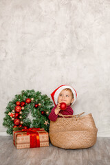 Sweet baby Santa sits in a wicker basket with a gift box and a Christmas wall on a gray background. Christmas concept, text space