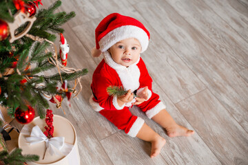 Cute baby Santa sits at home near the Christmas tree with gifts