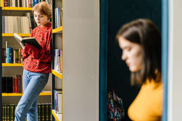 Adult attractive short hair woman in red sweater reading book while standing near bookshelves in...